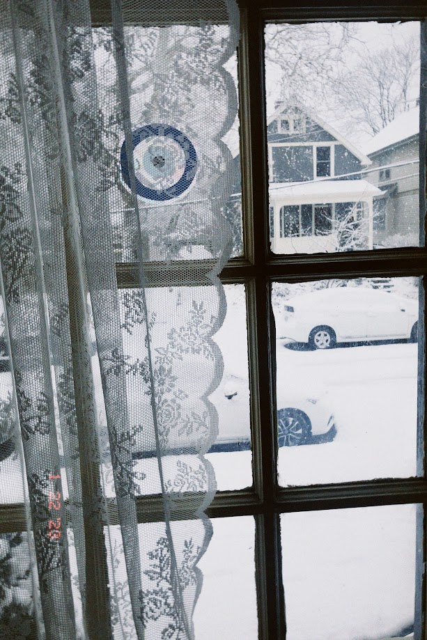 january 22, 2023: a snowy city street, as seen through a window with lace curtains and an evil eye suncatcher.
