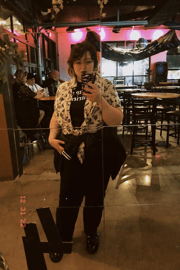 december 31, 2022: a mirror selfie of jude in a bar, wearing black jeans and an off-white button up with a skull pattern.