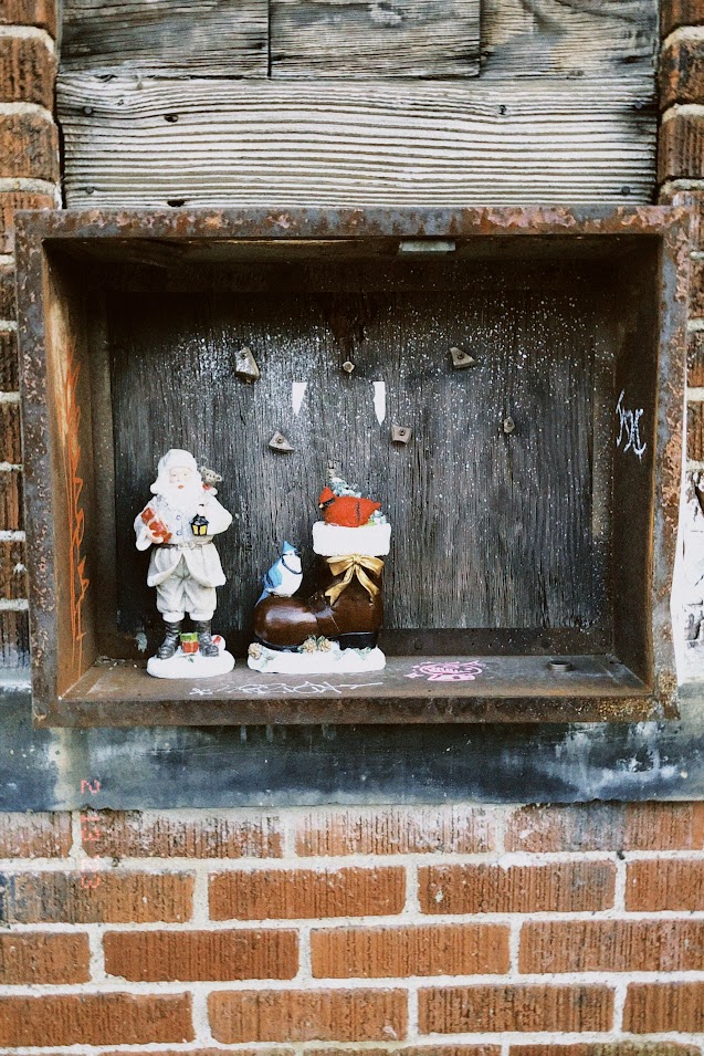 february 13, 2023: a ceramic Santa figurine and a ceramic boot with a cardinal and blue jay perched on it placed in a wooden shadowbox against a brick wall.