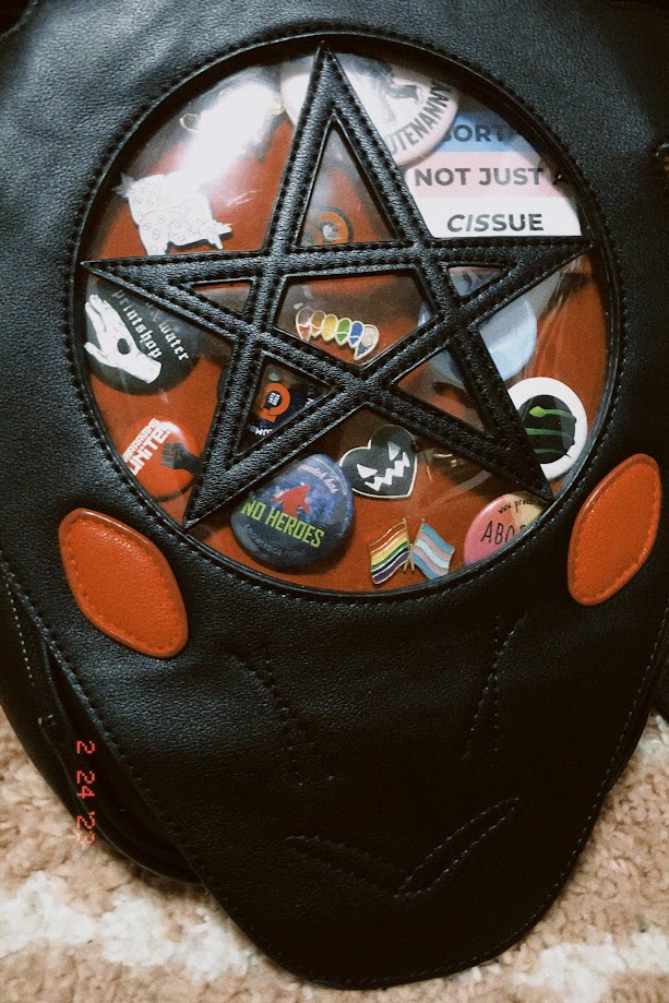 february 24, 2023: a black ita bag shaped like a goat with a pentagram on its forehead. the window of the bag is filled with a variety of pins and buttons.