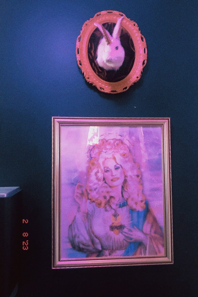 february 8, 2023: a gold-framed portrait of Dolly Parton as a saint hanging on a teal blue wall. above it is a taxidermied white rabbit with a unicorn horn.